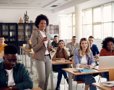 Professora sorridente apresentando conteúdo para uma turma de jovens adultos em uma sala de aula iluminada e moderna, com alunos atentos e engajados.